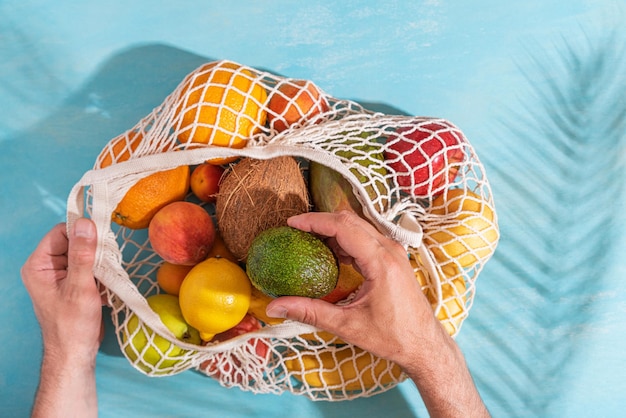 Polso dell'uomo che prende il frutto dell'avocado dalla borsa della spesa in rete con frutti tropicali Sfondo blu con ombra di foglia di palma