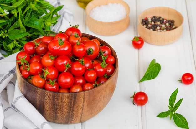 Pomodori ciliegia, erbe del basilico, olio d'oliva e sale dentro su una tavola di legno bianca della ciotola di legno