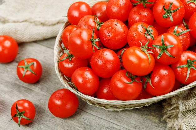 Pomodori ciliegia maturi e succosi con gocce di umidità in un cestino di vimini. Vecchio tavolo di legno, attorno alla tela di tela