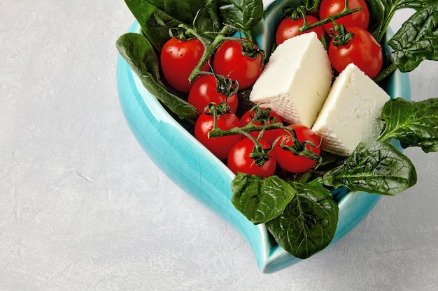 Pomodorini rossi su un ramo e foglie di spinaci verdi in una ciotola blu a forma di cuore su un tavolo di cemento chiaro