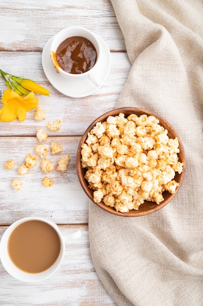 Popcorn con caramello in una ciotola di legno e una tazza di caffè su una superficie di legno bianca e tessuto di lino