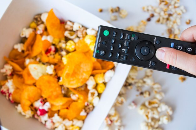 Popcorn fritto e patatine in una ciotola sullo sfondo bianco, mano che tiene il controller TV, vista ad angolo basso