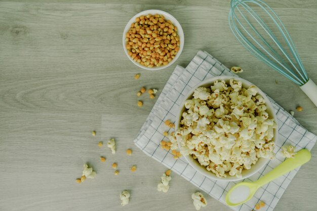 Popcorn in ciotola bianca con cucchiaio pastello, sale, miscelatore della mano e semi di mais su fondo di legno.