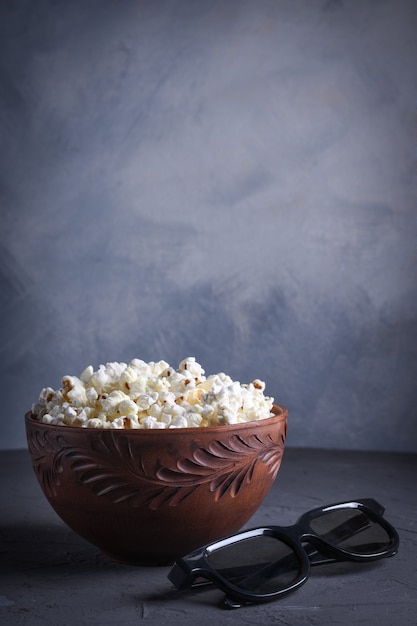 Popcorn salato in una ciotola con occhiali 3D su un tavolo su uno sfondo grigio. Vista frontale.