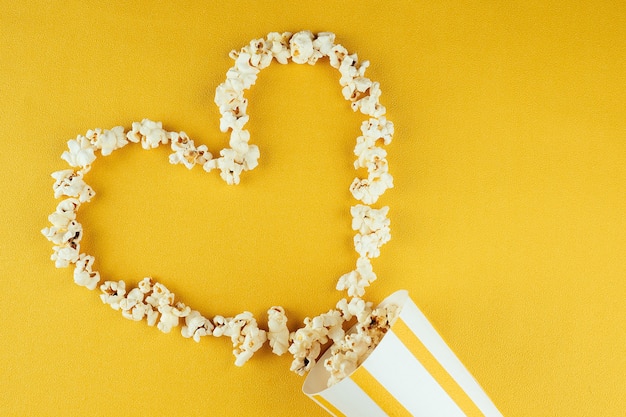 Popcorn sparsi da un bicchiere di carta a strisce su uno sfondo giallo a forma di cuore. il concetto di home cinema e film al cinema