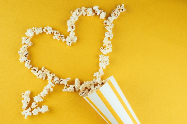 Popcorn sparsi da un bicchiere di carta a strisce su uno sfondo giallo a forma di cuore. il concetto di home cinema e film al cinema