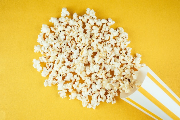Popcorn sparsi da un bicchiere di carta a strisce su uno sfondo giallo. il concetto di home cinema e film al cinema.