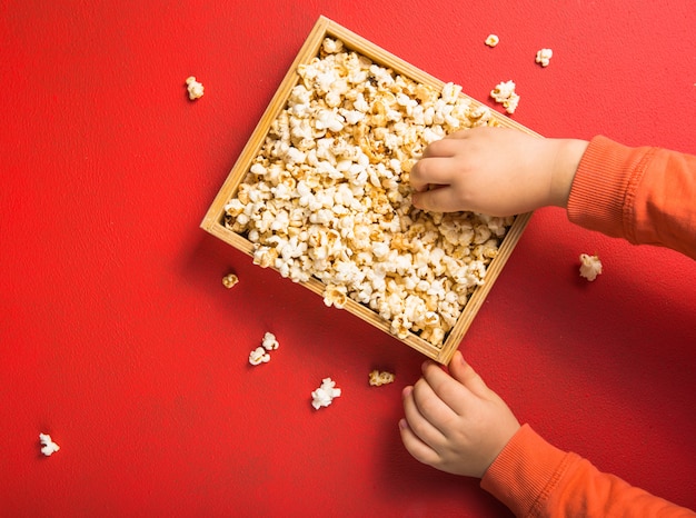 Popcorn visto dall'alto su sfondo rosso. Bambino che mangia popcorn. Mano umana. Concetto di snack cinematografici. Il cibo per guardare un film e divertirsi. Copi lo spazio per il testo, disteso.