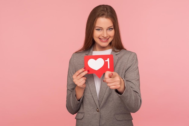 Popolarità sui social. Donna elegante sorridente in tailleur che tiene il cuore di rete Come l'icona e indica la fotocamera, consigliandoti di seguire il blog. girato in studio isolato su sfondo rosa