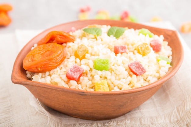 Porridge di bulgur con albicocche secche e frutta candita in ciotola di argilla su una superficie di cemento grigio e tessuto di lino. Vista laterale, messa a fuoco selettiva.