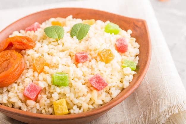 Porridge di bulgur con albicocche secche e frutta candita in ciotola di argilla su uno sfondo di cemento grigio e tessuto di lino. Vista laterale, messa a fuoco selettiva.