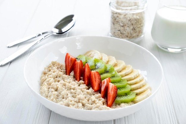 Porridge di farina d'avena con frutta fresca fragola banana kiwi in una ciotola bianca con bicchiere di latte vasetto con avena e cucchiaio sullo sfondo Ricetta di una sana colazione mangiare pulito