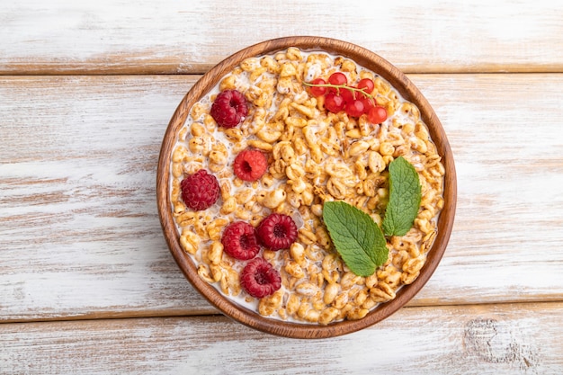 Porridge di fiocchi di grano con latte, lampone e ribes in ciotola di legno su superficie di legno bianca