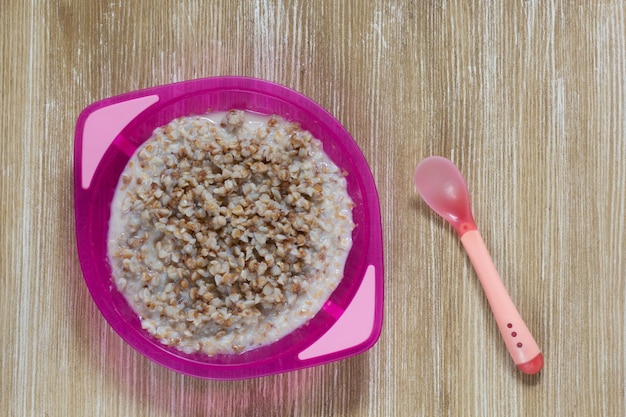 Porridge di grano saraceno con latte sul piatto di plastica rosso del bambino e cucchiaio su fondo di legno