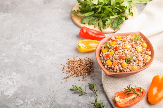 Porridge di grano saraceno con verdure in ciotola di argilla su una superficie di cemento grigio e tessuto di lino. Vista laterale, copia spazio.