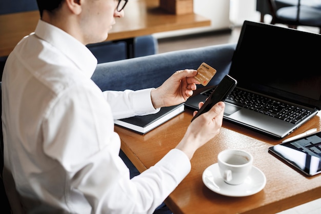 Porta carte di credito in oro da un uomo mentre si tiene e uno smartphone seduto a una scrivania con un laptop su di esso.