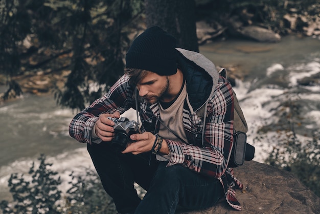 Porta sempre con te la macchina fotografica. Giovane uomo moderno con zaino che tiene in mano una macchina fotografica mentre è seduto nei boschi con il fiume sullo sfondo