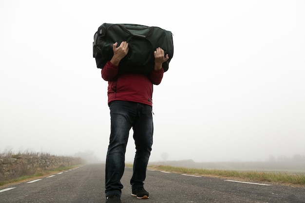 Portando una valigia tra le braccia un uomo irriconoscibile che cammina lungo una strada in una giornata nebbiosa