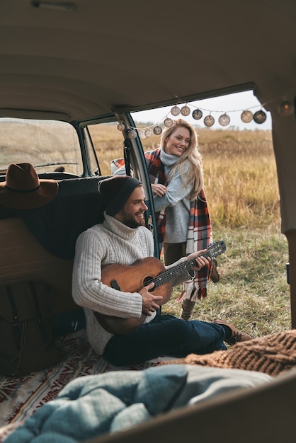 Portare musica ovunque. Bel giovane che suona la chitarra per la sua bella ragazza mentre è seduto in mini van in stile retrò