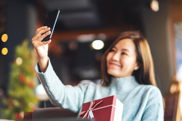 Portrait image of a beautiful young asian woman using mobile phone to take selfie with gift box