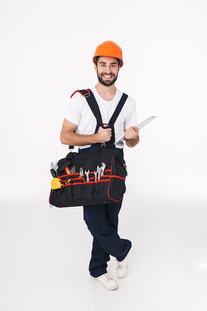 Portrait of cheerful smiling young man builder in helmet isolated over white wall holding bag with equipment instruments.