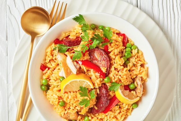 Porzione di paella di pollo spagnola con riso bomba valenciano, carne di coscia di pollo, salsicce chorizo, verdure e spezie servite su un piatto bianco su un tavolo di legno bianco, piatto, macro