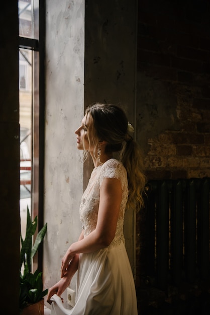 Posa di modello in un vestito da sposa lungo bianco all'interno dalla finestra