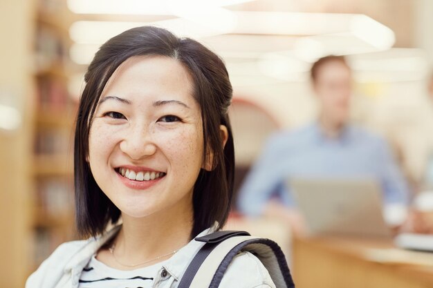 Positiva bella studentessa asiatica con i capelli neri corti in piedi in biblioteca e sorridendo alla telecamera