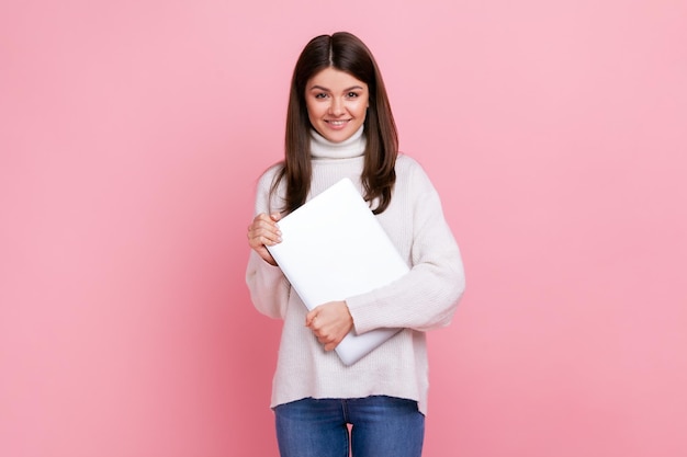 Positiva donna bruna ottimista in piedi con il laptop in mano, libero professionista pronto a lavorare, indossa un maglione bianco in stile casual. Studio indoor girato isolato su sfondo rosa.