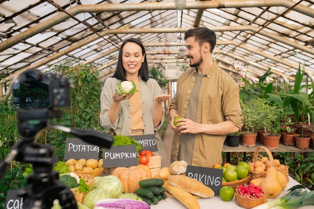 Positiva giovane coppia di agricoltori che parlano dei benefici del cavolfiore durante le riprese di video sulle verdure in serra