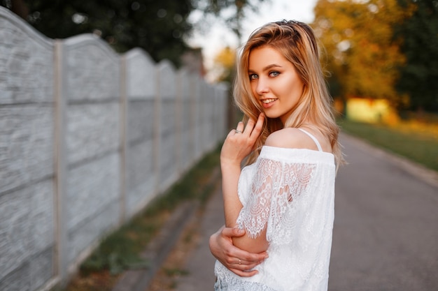 Positiva giovane donna attraente in un'elegante camicetta di pizzo in posa all'aperto vicino al recinto in una soleggiata giornata estiva