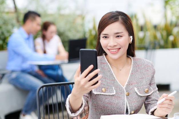 Positiva giovane imprenditrice vietnamita seduta al tavolo in caffè all'aperto e avendo videochiamata con un collega