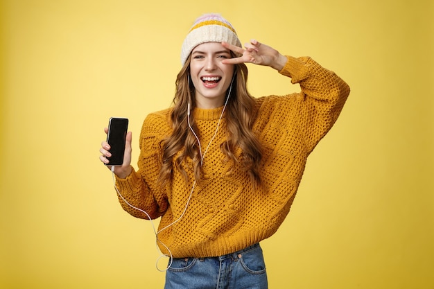 Positiva spensierata affascinante ragazza mostra gesto di pace indossando auricolari cablati che mostrano lo schermo dello smartphone che promuove l'app cool nuovissimo telefono cellulare, ridendo spensierato sfondo giallo