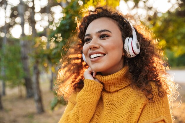 positivo allegro bella giovane donna riccia che cammina nel parco all'aperto ascoltando musica con le cuffie.