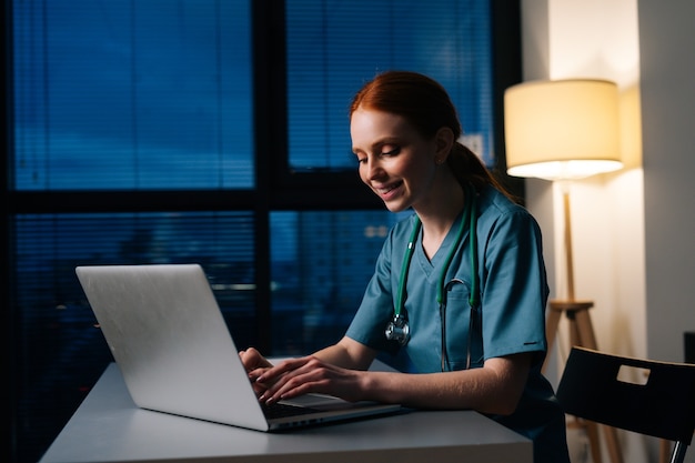 Positivo allegro rossa giovane dottoressa in uniforme medica verde blu che lavora digitando sul computer portatile seduto alla scrivania nella stanza buia dell'ufficio dell'ospedale vicino alla finestra di notte.