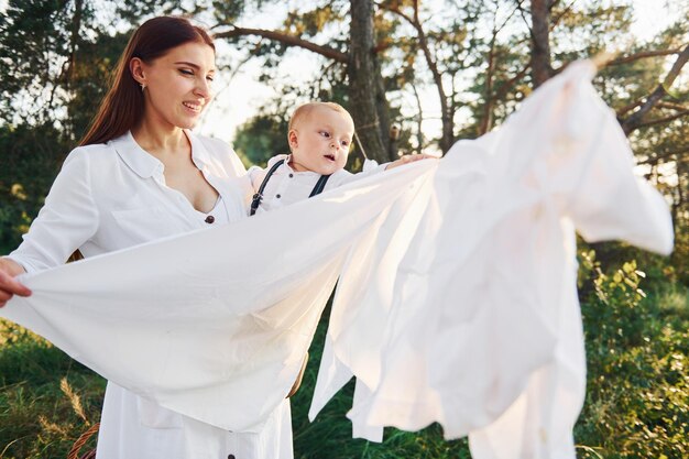 Positivo casalinga con appende abiti bianchi ad asciugare Giovane madre con il suo figlioletto è all'aperto nella foresta Bel sole