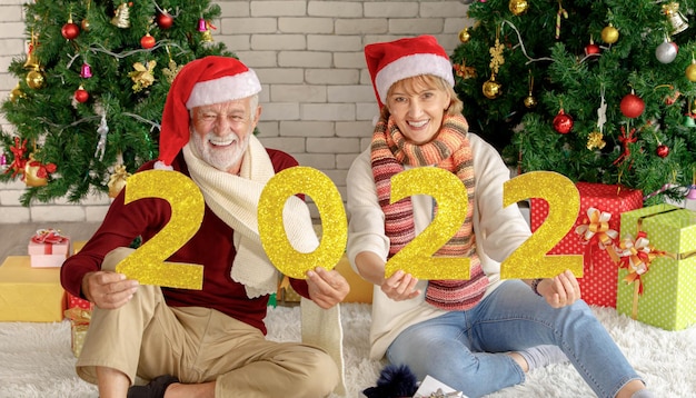 Positivo coppia senior guardando la fotocamera con un sorriso dimostrando golden numero 2022 contro un muro di mattoni un alberi di Natale durante la celebrazione del nuovo anno