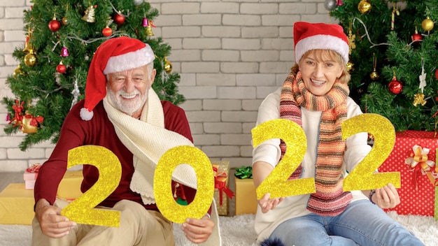 Positivo coppia senior guardando la fotocamera con un sorriso dimostrando golden numero 2022 contro un muro di mattoni un alberi di Natale durante la celebrazione del nuovo anno