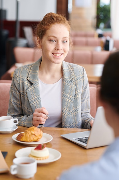 Positivo fiducioso giovane femmina specialista di marketing con i capelli rossi seduto al tavolo nella caffetteria e spiegando la strategia pubblicitaria al cliente alla riunione informale