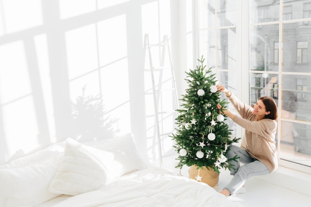 Positivo giovane femmina pone vicino alla grande finestra in camera da letto accogliente gode di decorare l'albero di Natale indossa abiti caldi ha un sorriso felice sul viso si prepara per la festa della famiglia celebrazione Felice Anno Nuovo