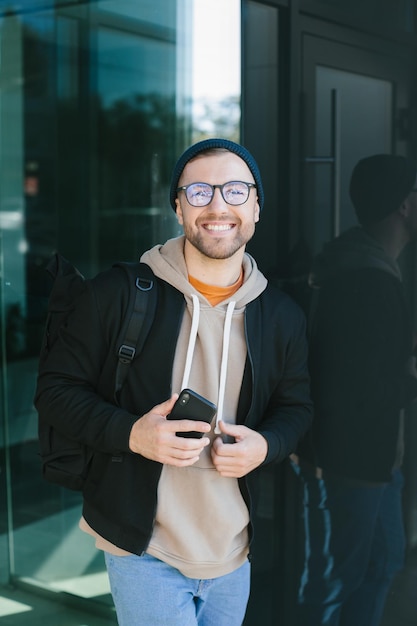 Positivo giovane ragazzo che tiene lo smartphone nelle sue mani guardando la fotocamera in piedi sulla strada Uomo barbuto in occhiali indossa abiti casual Concetto di utilizzo del telefono cellulare