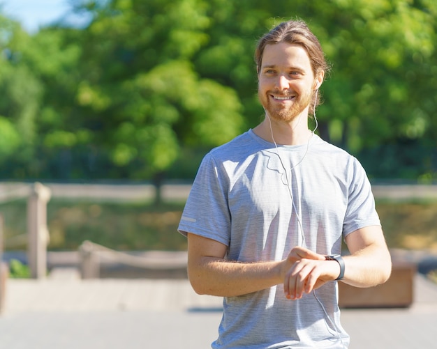 Positivo giovane sportivo bello in auricolari utilizzando il braccialetto fitness durante l'allenamento all'aperto