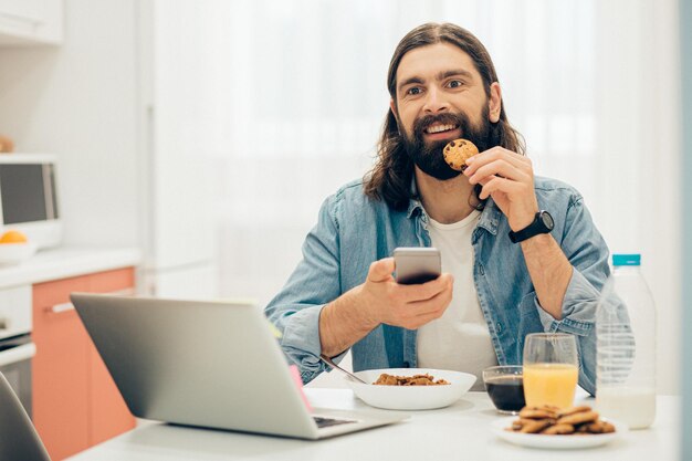 Positivo giovane uomo caucasico seduto al tavolo della cucina con un biscotto e uno smartphone in mano