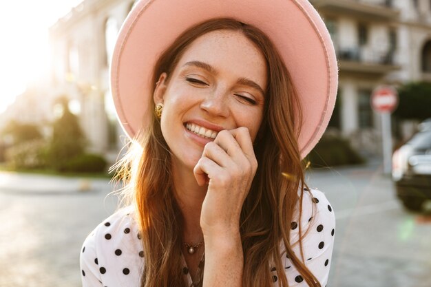 positivo sorridente ottimista felice giovane donna rossa che cammina per strada all'aperto in abito e cappello.