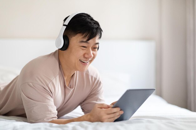 Positivo uomo coreano con tablet digitale e cuffie a letto