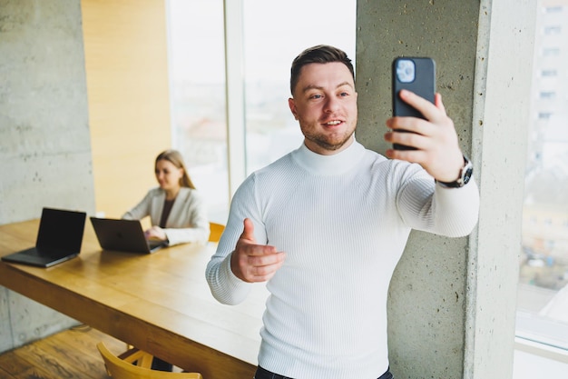 Positivo uomo d'affari maschio in maglione sorridente guardando il telefono in piedi in un moderno ufficio luminoso con telefono che lavora in un'azienda moderna