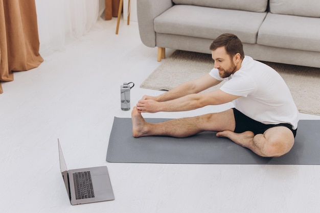 Positivo uomo di mezza età barbuto atletico che si allunga a casa utilizzando il computer portatile guardando video sportivi su Internet con spazio di copia online di lezioni di fitness Stile di vita sano sport in autoisolamento