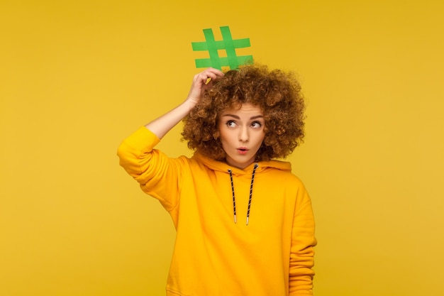 Post di blog popolari, contenuti alla moda. Ritratto di donna dai capelli ricci sorpresa in felpa con cappuccio in stile urbano che tiene un segno di hashtag sopra la testa, guardando con stupore. girato in studio isolato su sfondo giallo