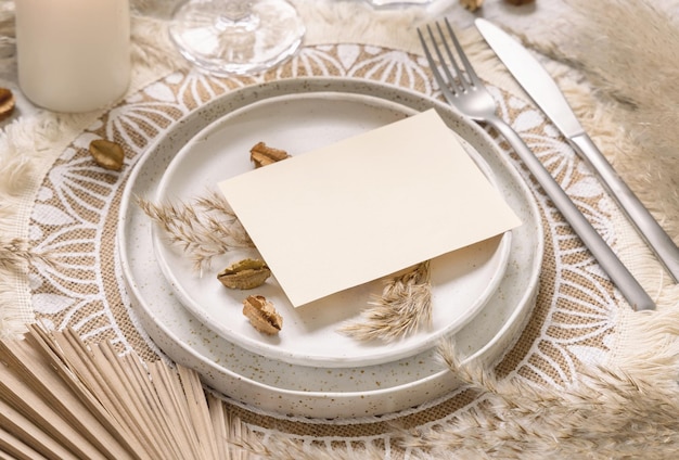 Posto del tavolo di nozze con carta in bianco orizzontale sulla tovaglietta bohémien da vicino. Modello di carta di matrimonio Boho. Modello beige tropicale con piante essiccate, foglie di palma ed erba di pampa, posto per il testo