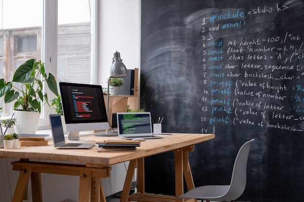 Posto di lavoro di esso ingegnere dalla finestra dell'ufficio con due laptop, monitor del computer, alcuni libri e quaderni, lampada e piante domestiche